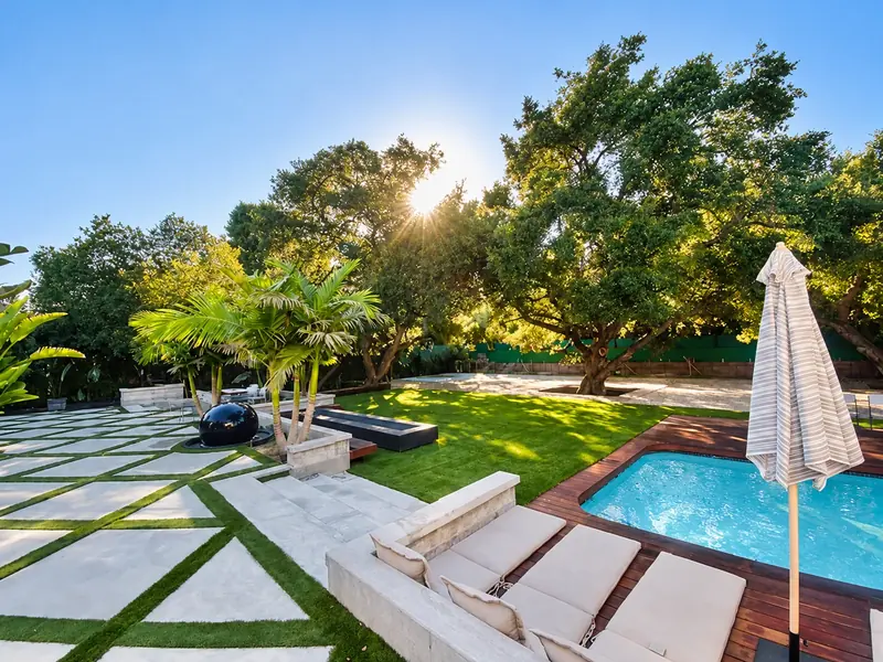 Poolside garden with geometric pale paving, green lawn, mature trees, a dark sphere fountain and swimming pool.