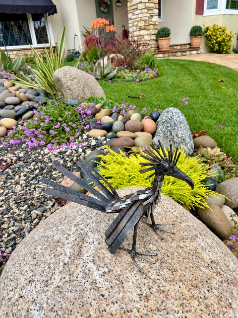 Metal bird sculpture on a boulder beside succulents, gravel, and rounded river stones.