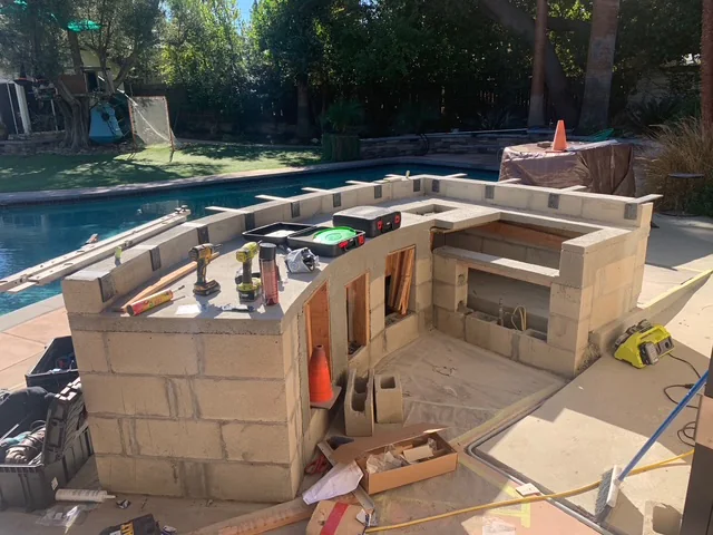 Curved masonry base for an outdoor BBQ island under construction beside a swimming pool.