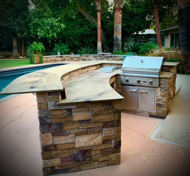 Curved outdoor BBQ island with stainless steel grill beside a kidney-shaped swimming pool and mature trees.