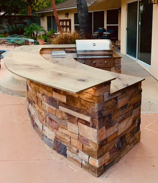 Custom curved BBQ island finished in earth-toned stacked stone beside the swimming pool.